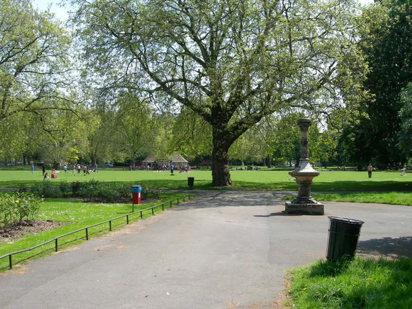 A wide path through Kennington Park leading toward a mature plane tree, with a Victorian ornamental fountain on the right and people on the grass in the middle distance