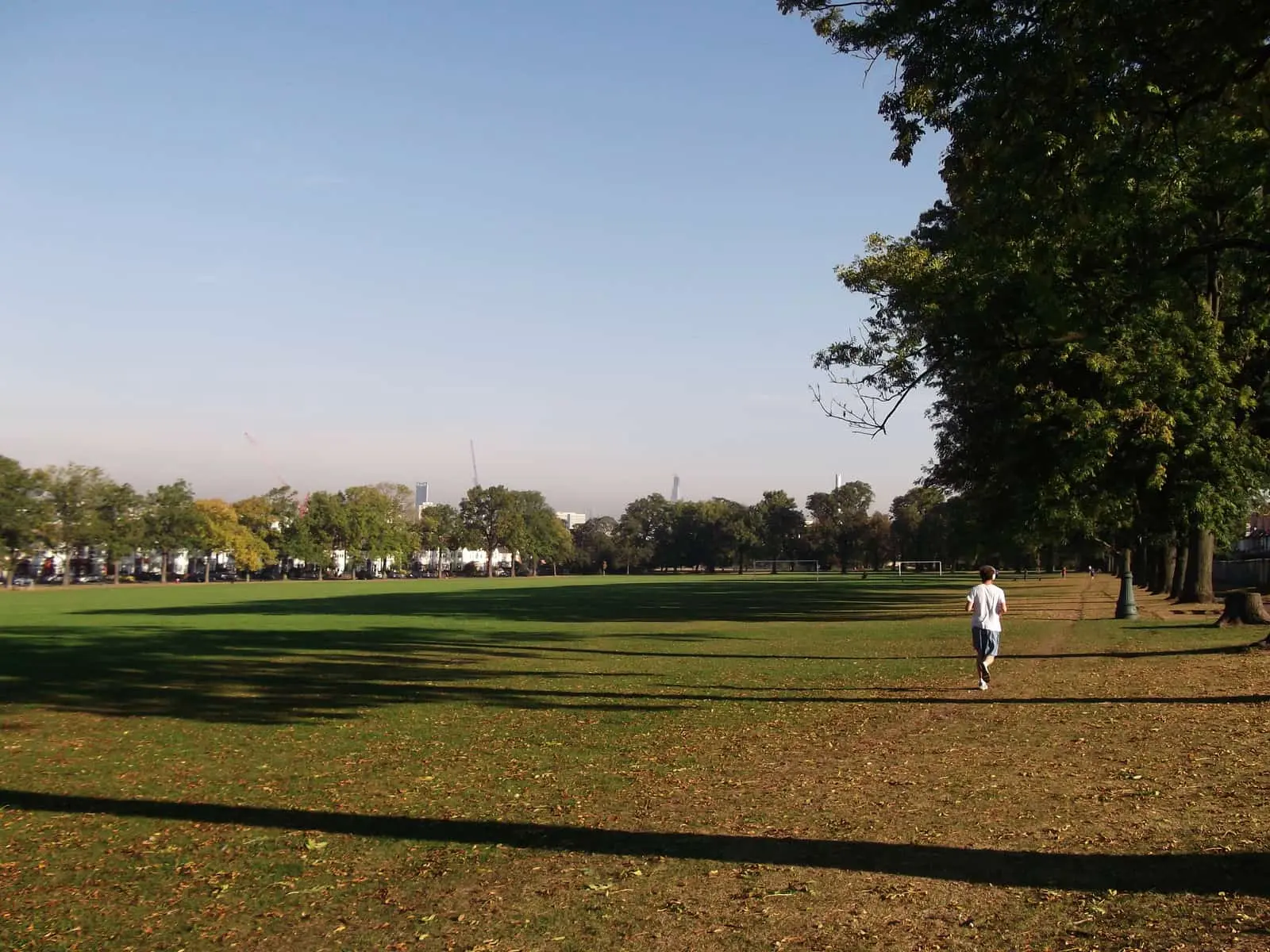 The lower playing fields of Ruskin Park on an autumn afternoon, with a jogger on the path and central London towers visible in the distance